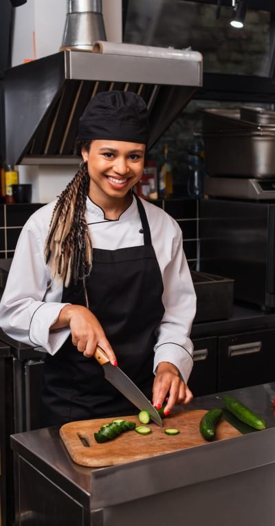 cheerful african american woman in apron cutting cucumber in modern kitchen